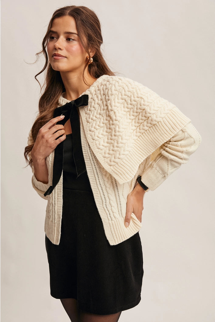 Woman wearing a beige knitted cardigan over a black dress on a plain background