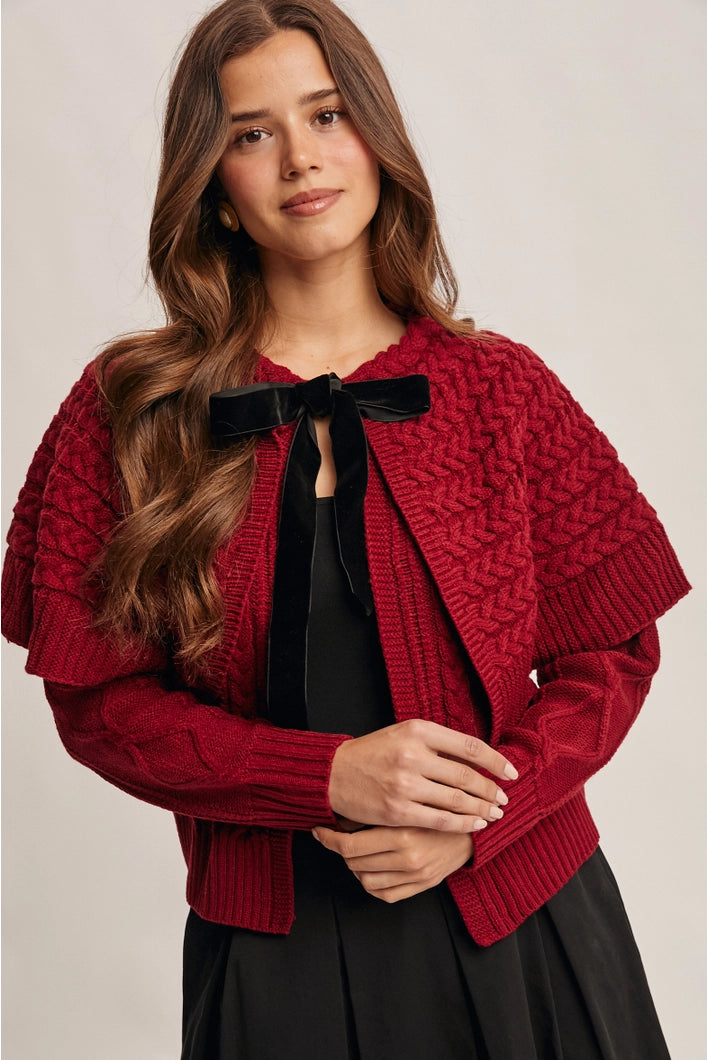 Woman wearing a red knitted cardigan with a black bow tie on a neutral background