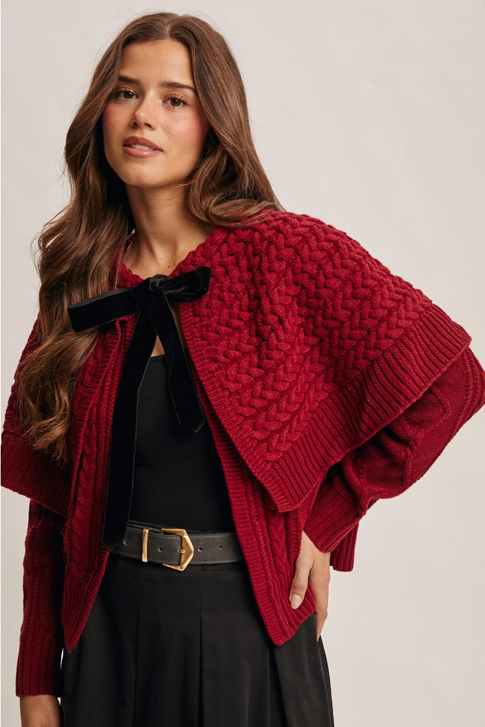 Woman wearing a red knitted poncho with a black bow on a plain background