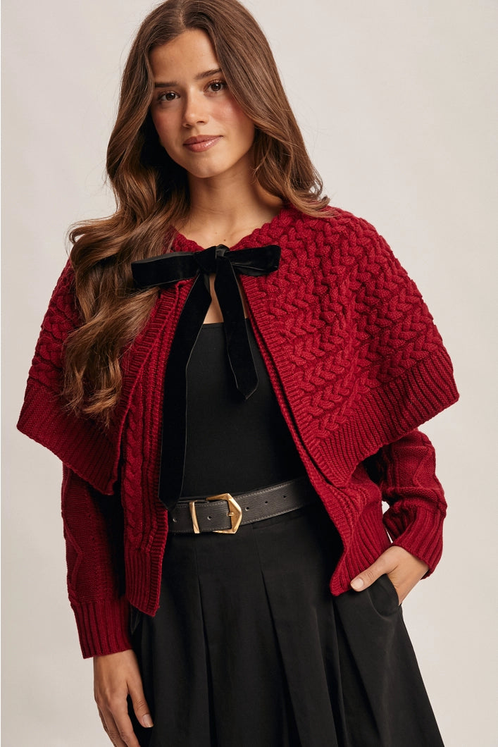 Woman wearing a red knitted cardigan with a black bow and black pants on a beige background