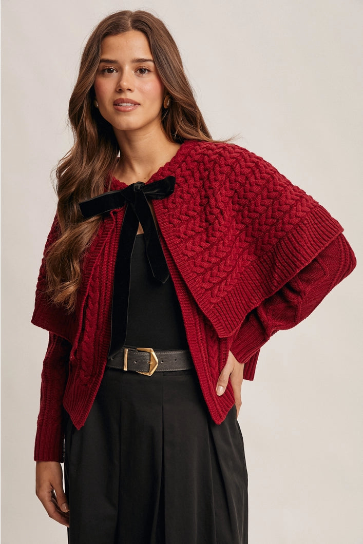 Woman wearing a red knitted poncho with a black bow and black pants on a beige background
