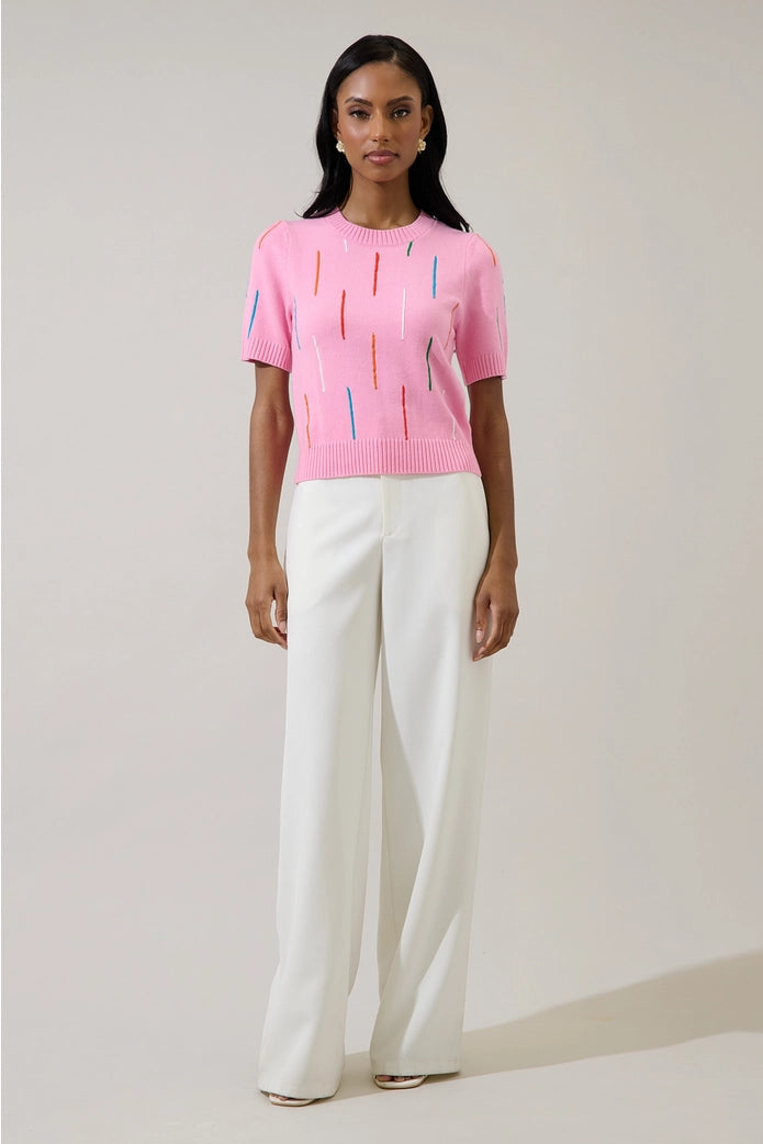 Woman wearing a pink knit top with colorful patterns and white pants against a plain background