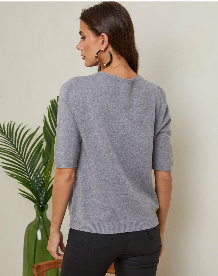 Woman wearing a gray sweater standing next to a plant against a plain background