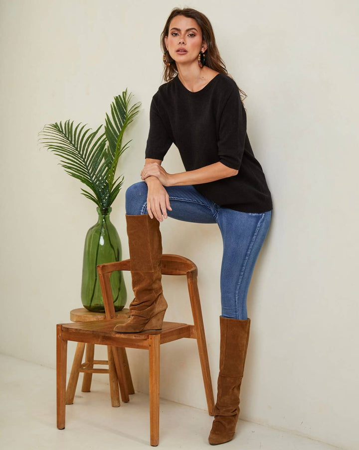 Woman wearing brown knee-high boots and blue jeans sitting on a wooden chair with a plant in the background.
