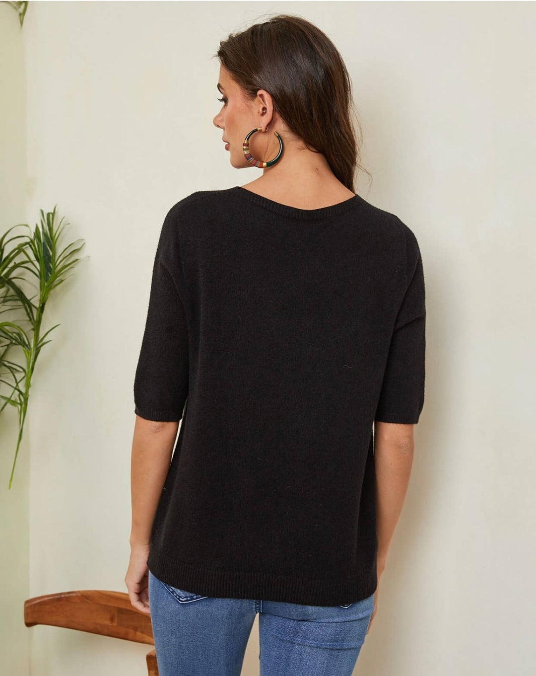 Woman wearing a black sweater and blue jeans against a white wall with a plant to the left.