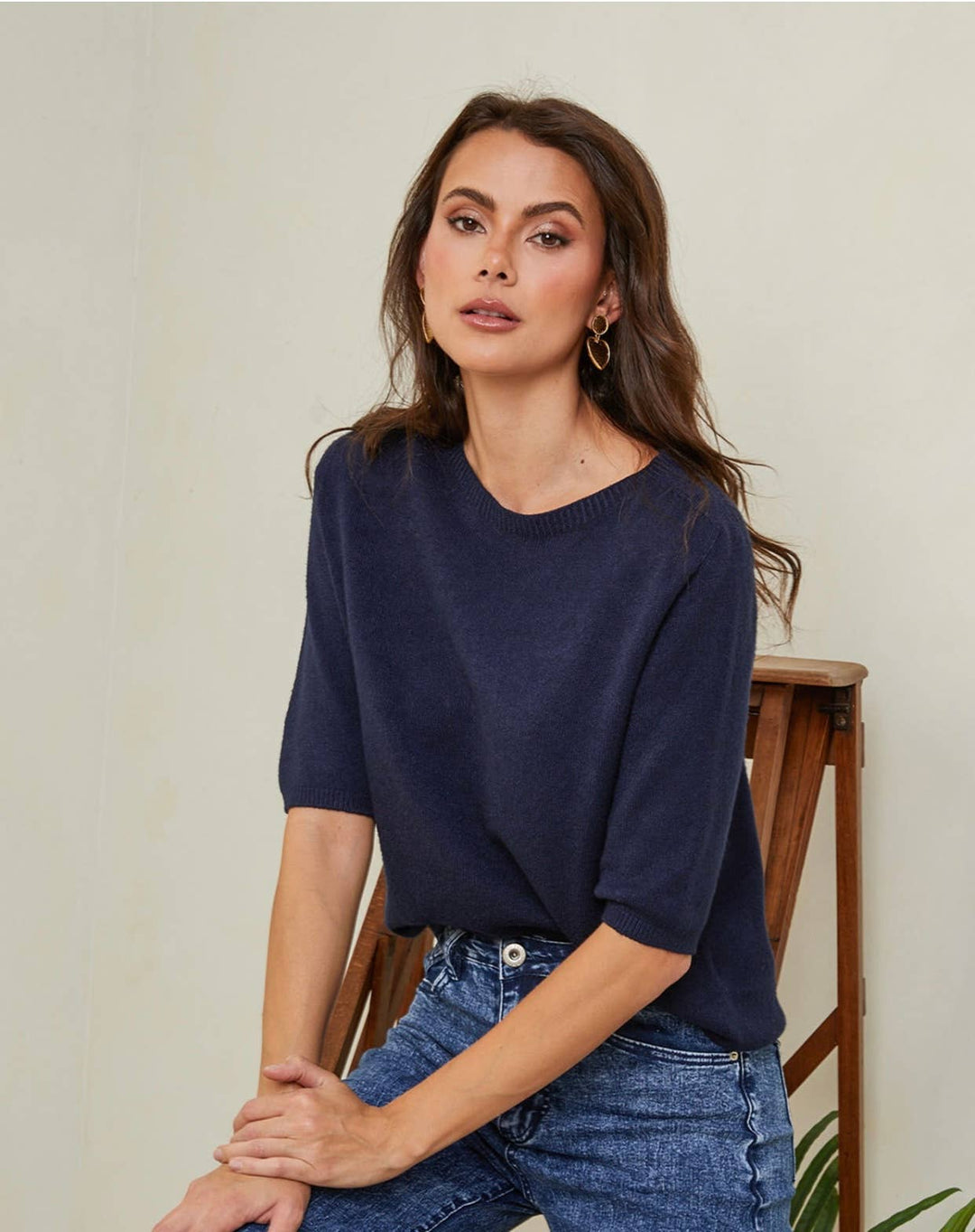 Woman wearing a navy blue top and jeans sitting on a wooden chair against a plain background