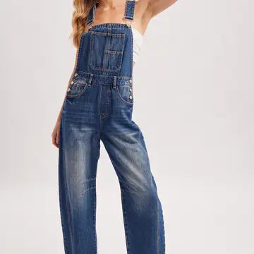 A model wearing blue denim wide-leg overalls with a faded wash, styled over a white tank top against a light gray studio background.