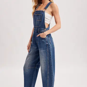 A model wearing blue denim overall jumpsuit with wide legs, a front bib pocket, and a side cutout, paired with a white ribbed tank top, against a light gray studio background.