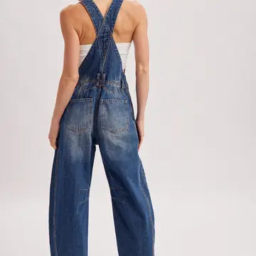 A model shown from the back wearing blue denim overalls with wide legs and crossed shoulder straps, photographed against a plain light gray studio background.