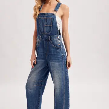 A model wearing blue denim overall-style pants with wide legs, front pockets, and decorative silver buttons on the bib, shown against a plain light gray studio background.