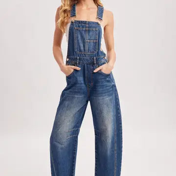 A model wearing blue denim overall-style jeans with a front bib pocket, shoulder straps, and wide legs, shown against a plain light gray studio background.