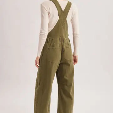 A person shown from the back wearing olive green bib overalls over a cream long-sleeve top, photographed against a plain light gray studio background.