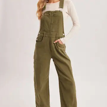 A model wearing olive green wide-leg denim overalls over a white long-sleeve top, shown from the waist up against a light gray studio background.