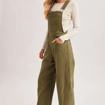 A model wearing olive green wide-leg denim overalls over a cream long-sleeve top, shown from the waist down against a light gray studio background.