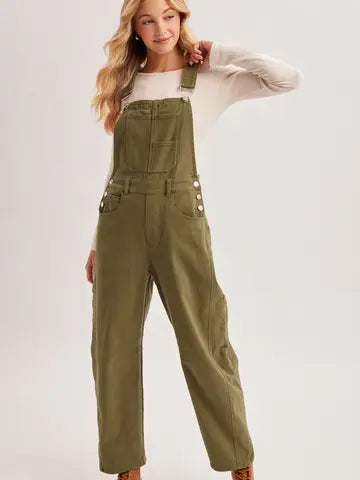 A woman modeling olive green wide-leg denim overalls over a white long-sleeve top, standing against a light gray studio background.