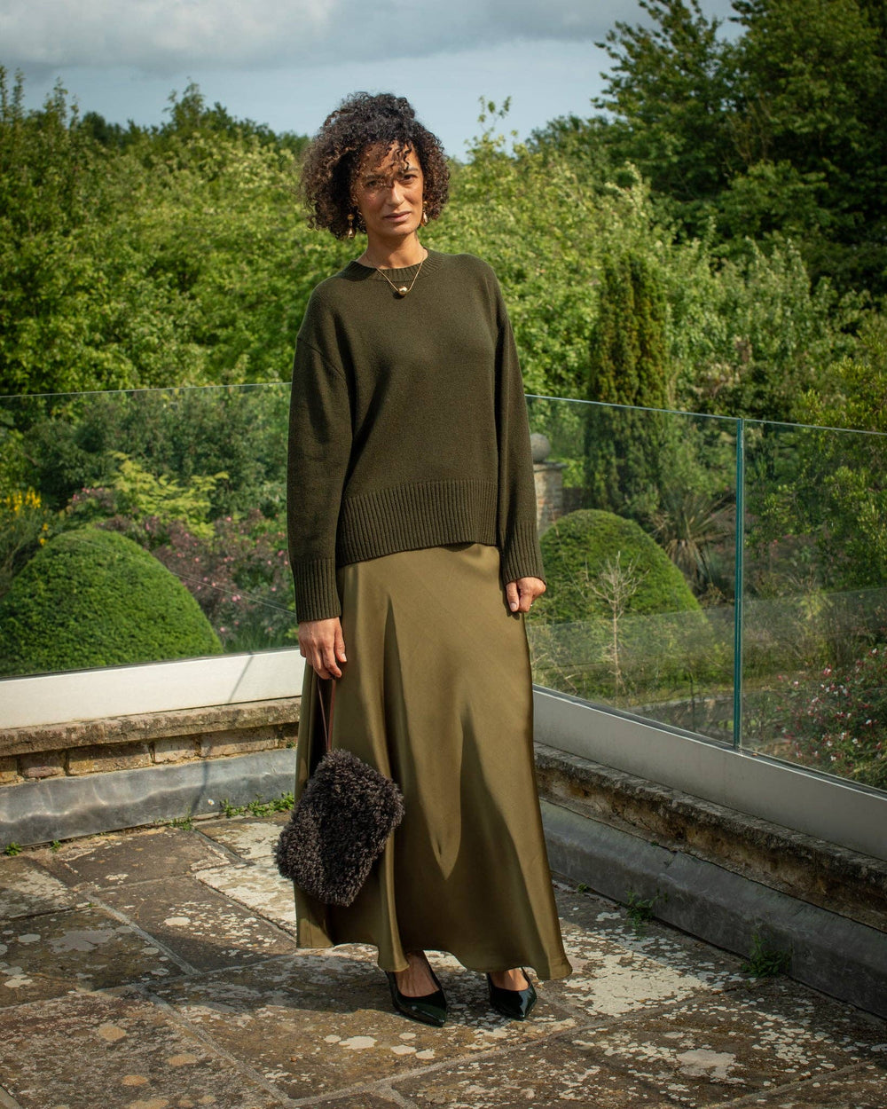 Woman in a green sweater and skirt standing outdoors with greenery in the background