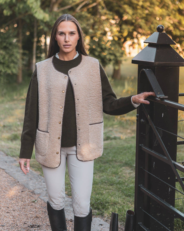 Woman wearing a beige vest over a dark sweater and white pants, standing next to a black gate with trees in the background.
