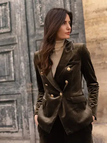 A woman wearing a dark olive green velvet double-breasted blazer with gold buttons over a beige turtleneck, standing in front of a rustic wooden door.