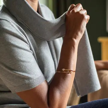 A close-up lifestyle shot of a person wearing a delicate gold bangle bracelet with a safety-pin style clasp, paired with a light gray knit sweater.