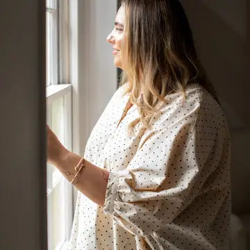 A woman in a cream-colored, long-sleeve button-up shirt with small black polka dots stands by a window, looking out and smiling. She has a gold bracelet on her wrist.
