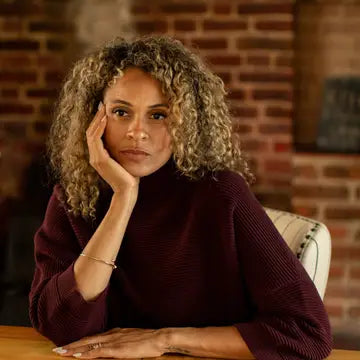 A woman with curly light brown hair rests her head on her hand, wearing a dark burgundy ribbed turtleneck sweater, seated in front of a brick wall background.