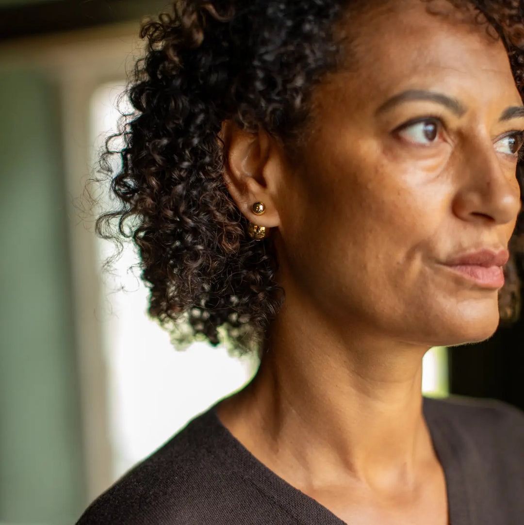 A close-up portrait of a woman with curly dark hair wearing a small gold hoop earring, shown in profile against a softly blurred indoor background.