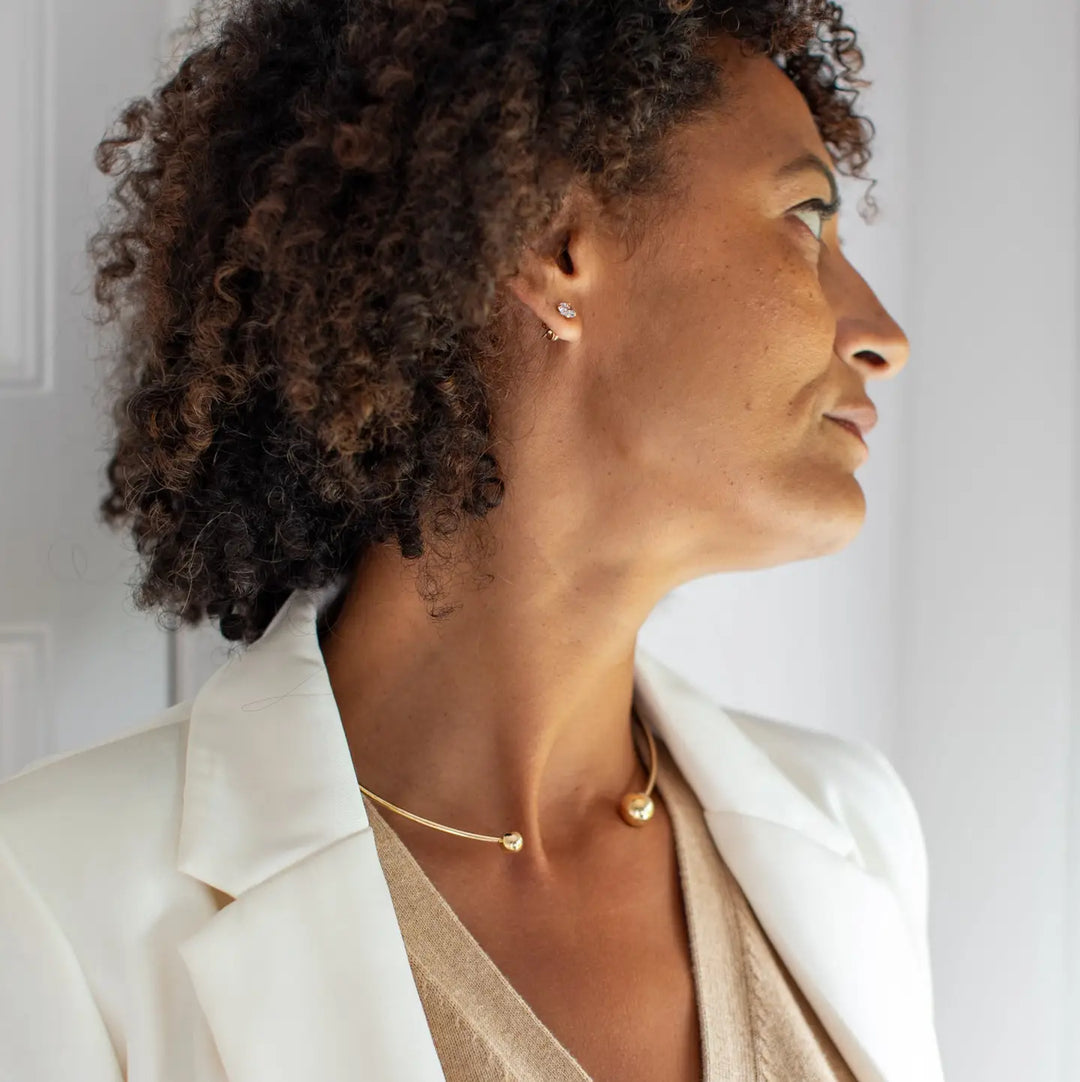 A woman in profile wearing a delicate gold necklace with a small gold bead pendant and a matching stud earring, styled with a white blazer and beige top.