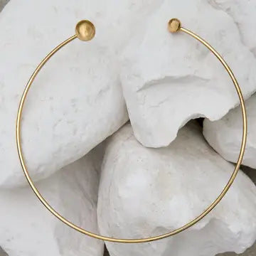 A minimalist gold hoop earring with a small spherical stud at each end, displayed on a textured background of white stones.