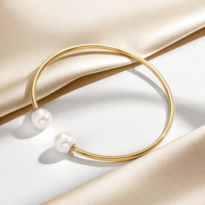A gold open cuff bracelet with two white pearl beads, displayed on a draped beige satin fabric background.