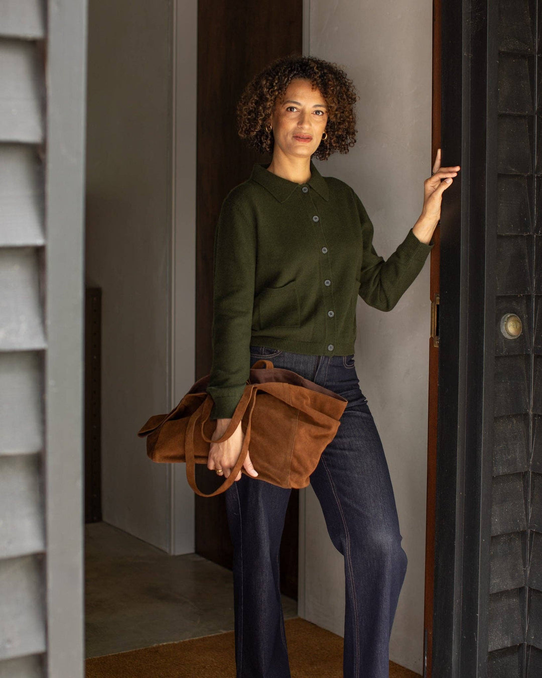 Woman holding a brown leather bag, wearing a green sweater and dark jeans, standing in a doorway.