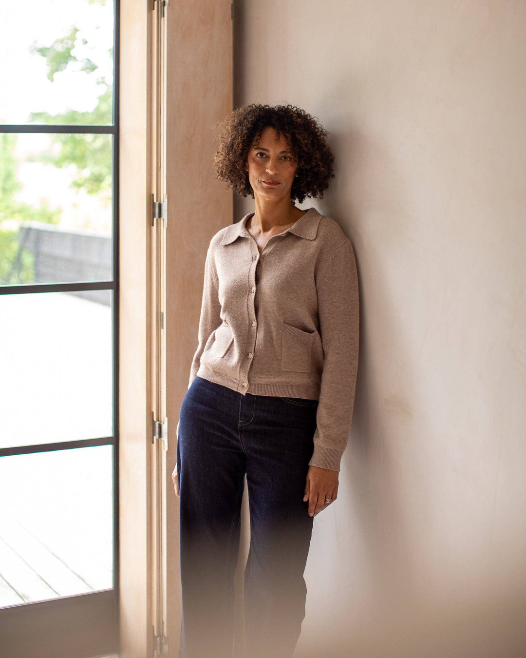 Woman leaning against a wall indoors with a window in the background