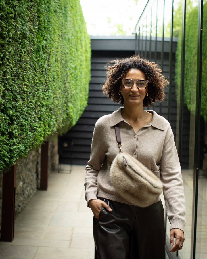 Woman with a beige sweater and black pants standing in an outdoor setting with greenery.