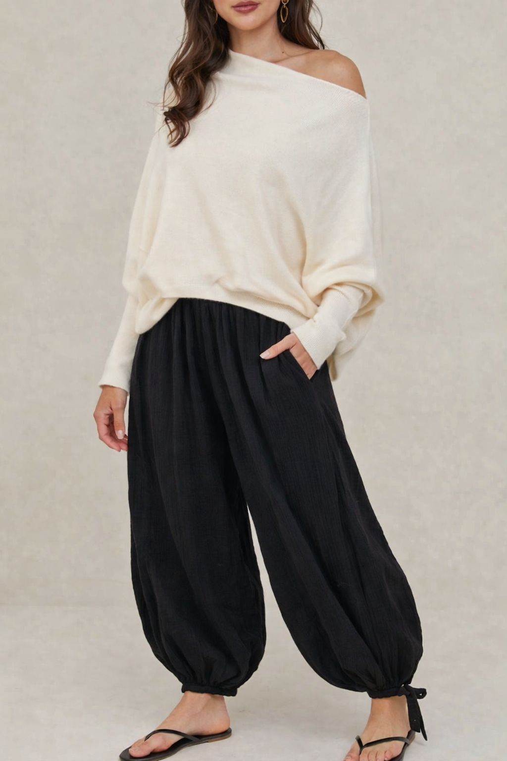 Woman wearing a white off-shoulder top and black harem pants on a plain background