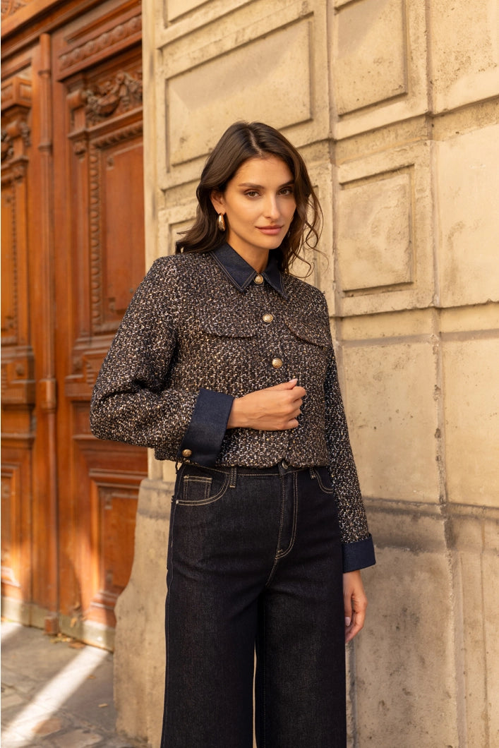 Woman wearing a patterned jacket and dark jeans standing against a textured wall.