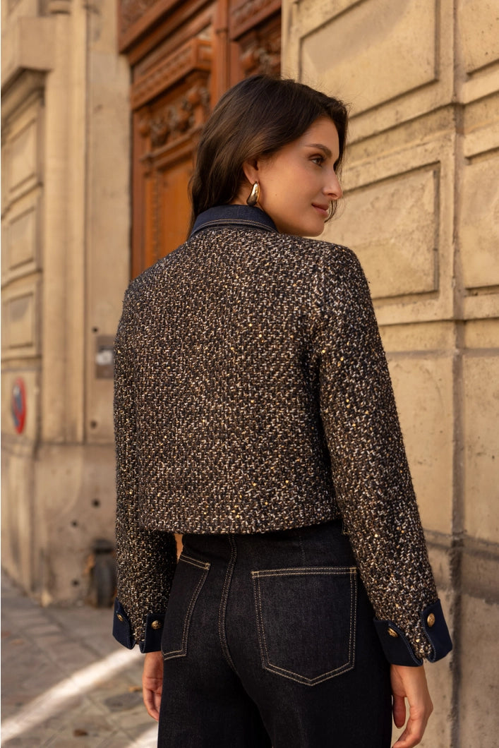 Woman wearing a patterned jacket and dark jeans standing against a textured wall.