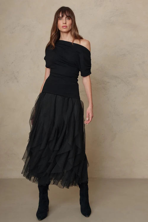 A woman modeling a black off-the-shoulder dress with a ruched bodice and a layered tulle midi skirt, paired with black ankle boots, against a textured beige studio backdrop.