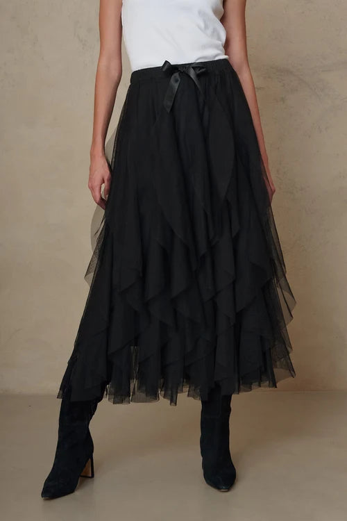 A model wearing a long black tiered tulle midi skirt with a matching black ribbon tie at the waist, styled with a white top and black ankle boots against a textured beige studio backdrop.
