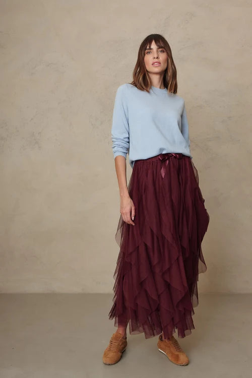 A woman stands against a textured beige studio wall wearing a light blue long-sleeve top, a long burgundy tulle skirt, and brown suede shoes.