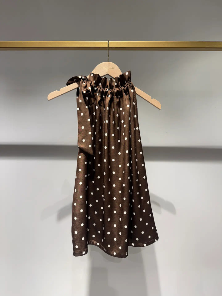 A sleeveless brown dress with white polka dots hanging on a wooden hanger against a light gray wall.