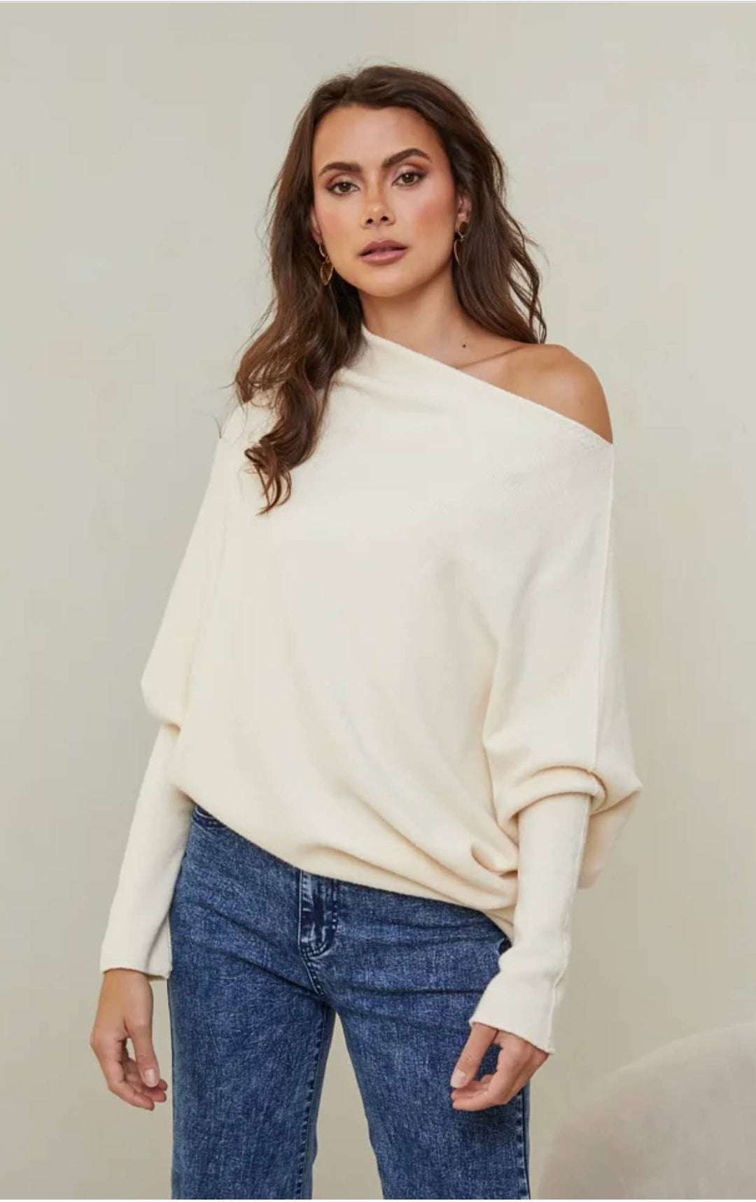 Woman wearing a white off-shoulder sweater and blue jeans against a plain background