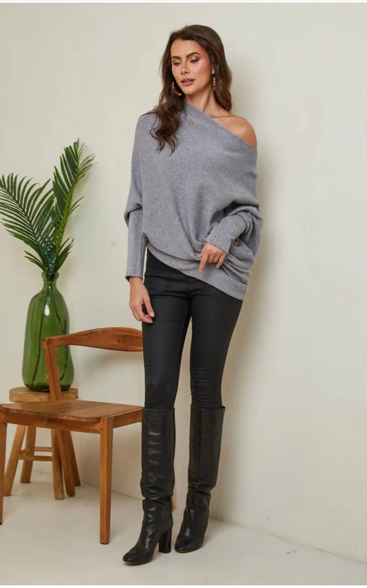 Woman wearing a gray off-shoulder sweater and black pants in a minimalistic room.