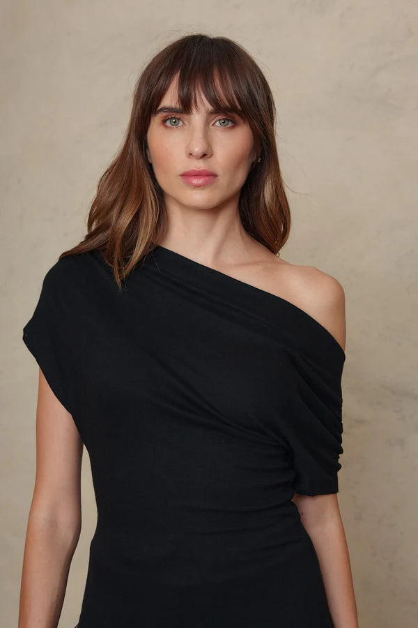 A woman with brown hair and bangs wearing a black draped one-shoulder dress, standing against a textured beige background.