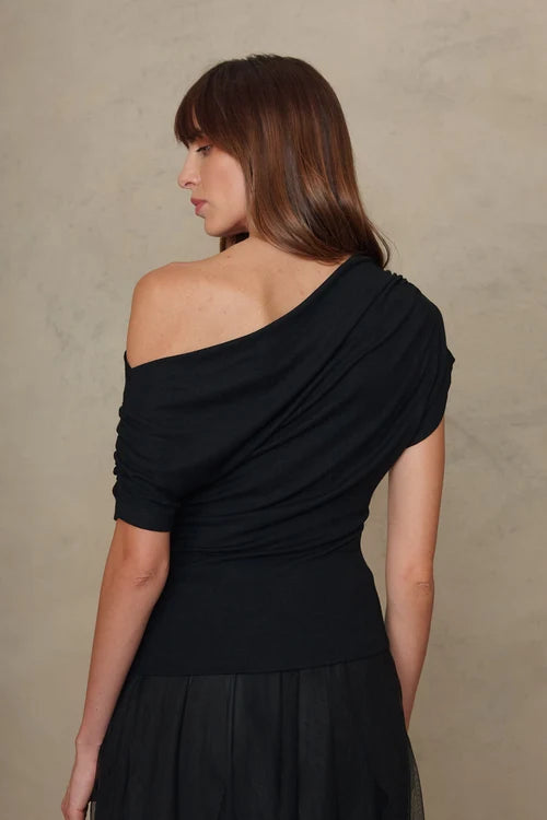 A woman shown from the back wearing a black draped one-shoulder top and matching black skirt against a textured beige studio backdrop.