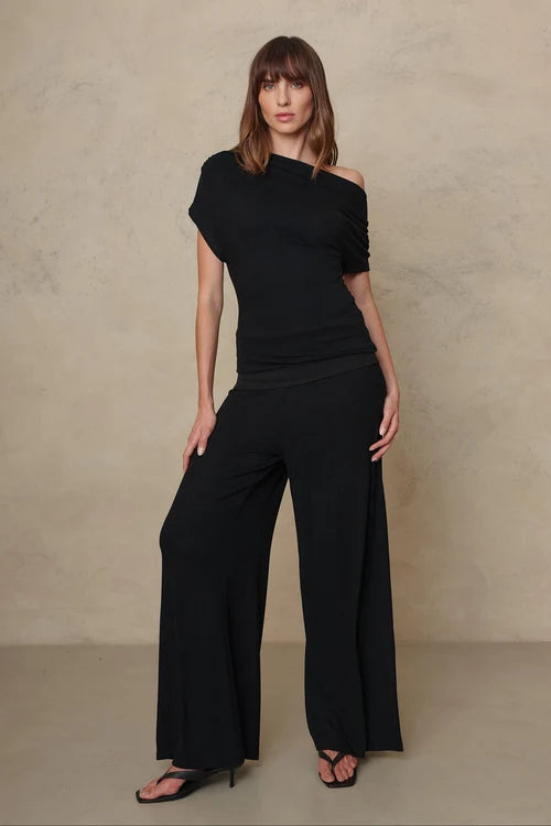 A woman modeling a black one-shoulder top and matching wide-leg trousers against a textured beige studio backdrop.