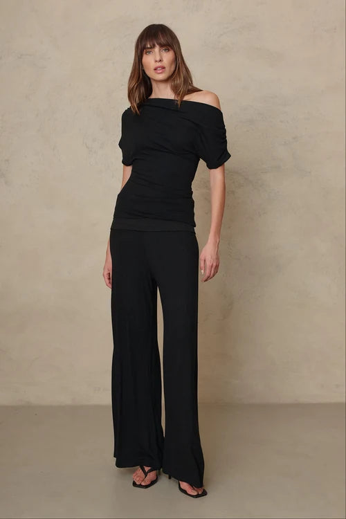 A woman modeling a black off-the-shoulder short-sleeve top paired with matching black wide-leg pants, standing against a textured beige studio backdrop.