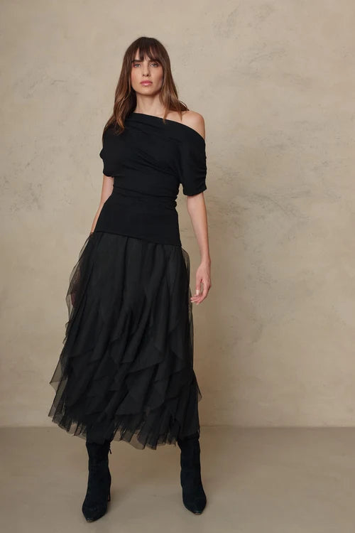 A woman modeling a black off-the-shoulder top with a matching layered tulle midi skirt and black ankle boots, standing against a textured beige studio backdrop.