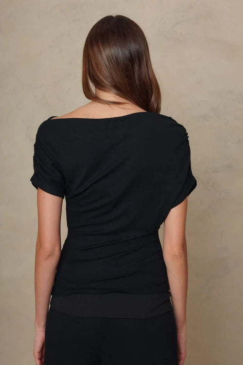 A woman viewed from the back wearing a black off-the-shoulder short-sleeve top against a textured beige studio backdrop.