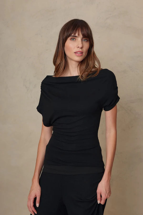 A woman modeling a black short-sleeve top with a draped, gathered neckline and ruched front, paired with black pants, against a textured beige studio backdrop.