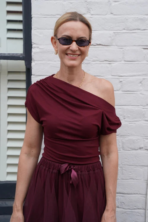 A woman wearing a burgundy one-shoulder top and matching skirt stands in front of a white brick wall.
