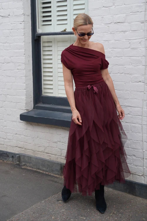 A woman wearing a burgundy off-the-shoulder midi dress with a tiered tulle skirt, styled with black ankle boots and sunglasses, standing on a sidewalk in front of a white brick wall.